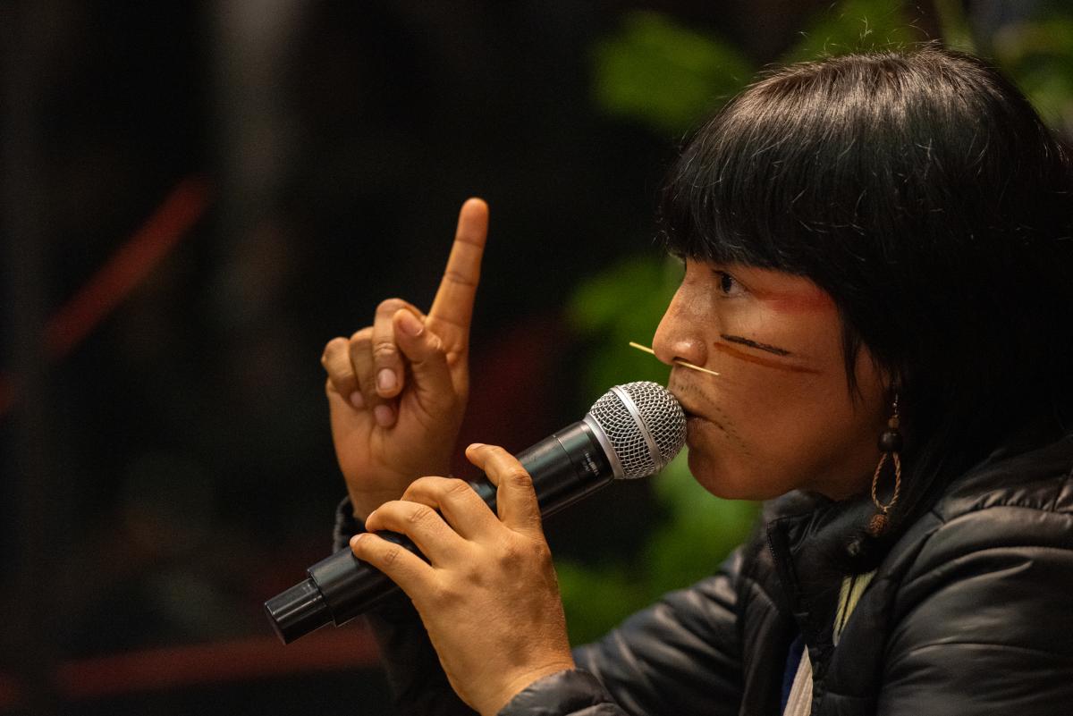 Ehuana Yanomami aparece de lado, com o dedo indicador para cima. Ela fala ao microfone