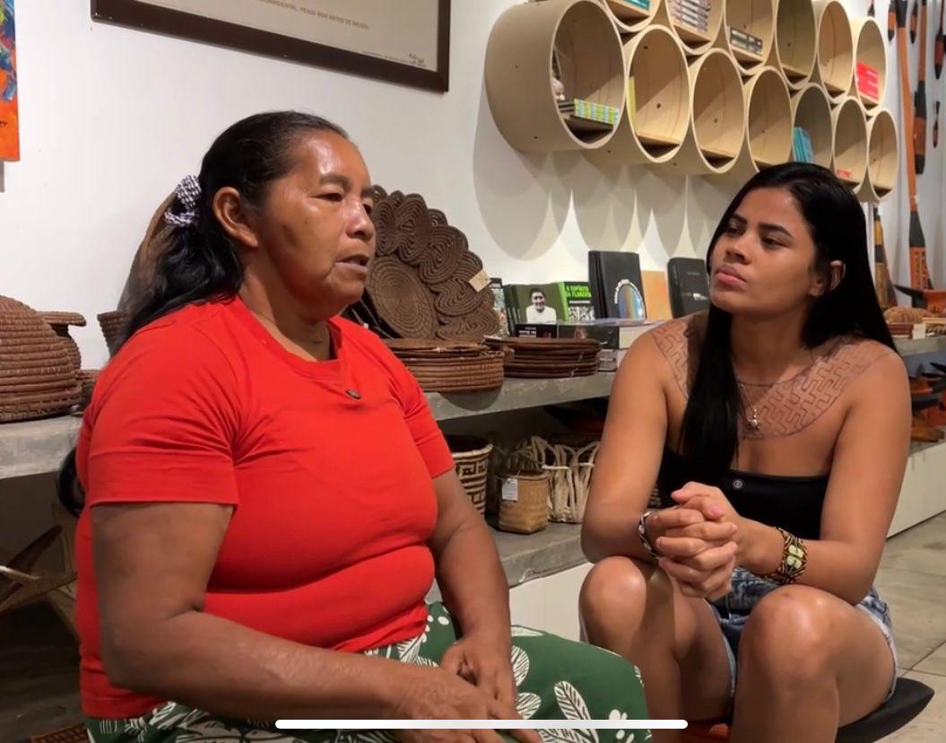 Imagem mostra uma senhora dando entrevista para uma jovem. Ambas estão sentadas e atentas uma a outra